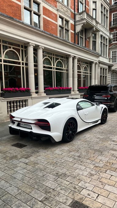 A white sports car parked on a cobblestone street in front of an elegant brick building with white columns and flower boxes, with a dark SUV visible behind it.
