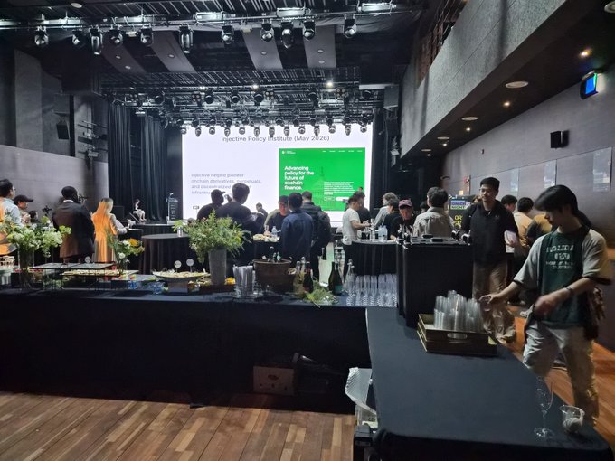 A crowded indoor event venue with stage lighting and a presentation screen reading 'Injective Policy Institute (May 2026)' and 'Advancing policy for the future of finance', with people mingling around black tables holding drinks and refreshments.