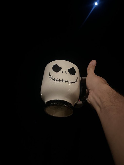 A Jack Skellington-themed ceramic mug shaped like the character's head from 'The Nightmare Before Christmas' is held up against a dark background by a person's hand.