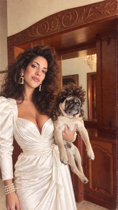 A woman with voluminous curly dark hair wearing a white satin dress with a deep neckline, holding a small tan pug, standing in front of an ornate wooden doorway with a chandelier visible in the background.