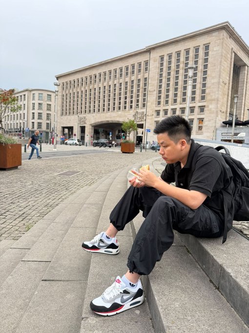 A young man wearing black clothing, a backpack, and white Nike sneakers sits on stone steps eating a snack, with a large classical building in the background.