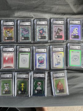 A collection of graded VeeFriends trading cards displayed in plastic slabs, arranged in three rows on a dark surface. The slabs feature various character designs including a yellow bird, purple creature, gorilla, and others, with numerical ratings and labels visible on each card holder.