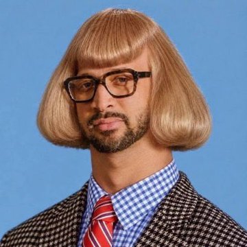 A portrait of a bearded man wearing thick black-framed glasses, a blonde bowl-cut wig, a blue checkered shirt, red striped tie, and a checkered blazer against a solid blue background.