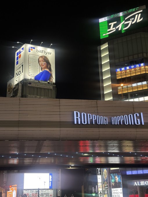 A nighttime view of the Roppongi station entrance illuminated with its signature "ROPPONGI" sign, with a bitFlyer billboard displayed on the building above, supporting the post's claim about bitFlyer's presence in Roppongi. Additional visible signage includes Japanese text "エイブル" (Able) and various storefronts, reinforcing the urban Tokyo setting.