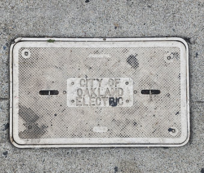 A photograph of a worn metal utility cover on a sidewalk that reads 'CITY OF OAKLAND ELECTRIC', with the letters 'BNC' spray-painted in black next to it.
