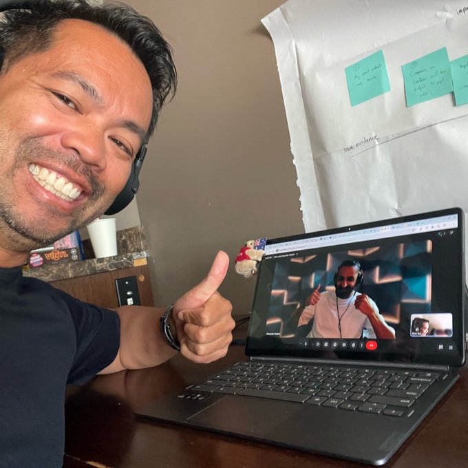 A smiling man wearing a headset gives a thumbs-up while on a video call displayed on his laptop, with sticky notes visible on the wall behind him.