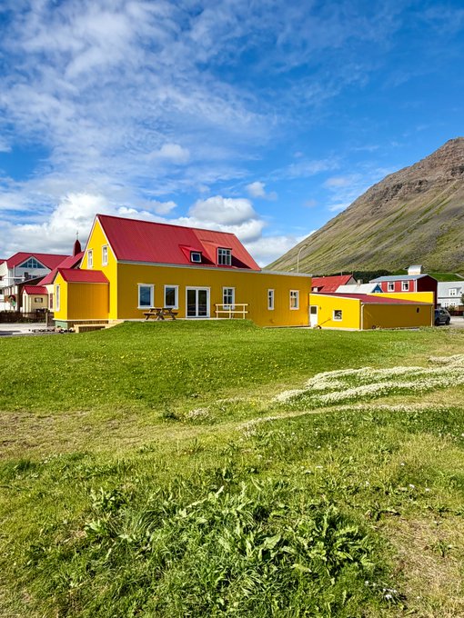 A bright yellow house with a red roof sits on a grassy hill under a blue sky, with other colorful houses and a mountain in the background.