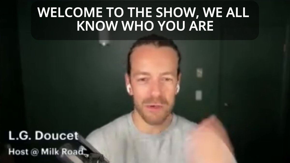 The image features a man wearing a grey shirt, standing in front of a green wall with text displayed above him. The text reads "Welcome To The Show, We All Know Who You Are", which seems to be a promotional message for a show or event. The man is also holding a cell phone and appears to be engaged in a conversation on it.
