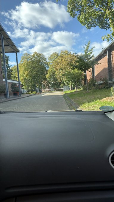 A view from inside a car looking out over the dashboard through the windshield, showing a driveway lined with trees and brick buildings under a partly cloudy blue sky.