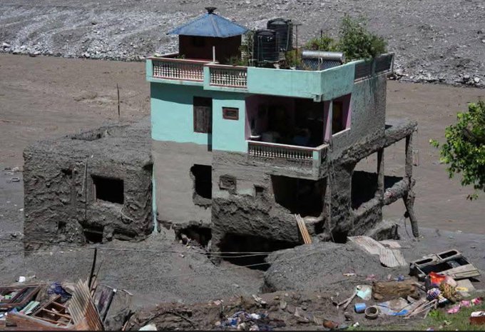 A photograph showing a partially constructed or damaged multi-story building with a blue-roofed top floor, surrounded by a flood or mudslide, set against a rocky, barren background.