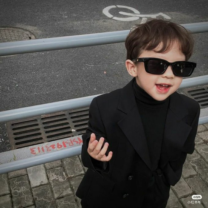 A young boy wearing sunglasses and a black blazer over a turtleneck poses on a street with a metal railing, with a "小红书号" (Xiaohongshu) watermark in the corner.