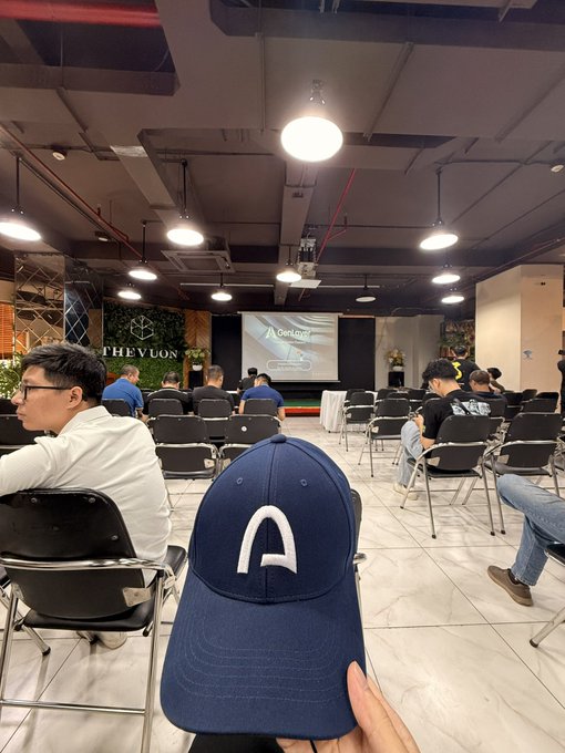 A first-person perspective of someone holding a navy blue cap with a white 'A' logo at an indoor event, with attendees seated in chairs facing a projector screen at THE VUON venue.