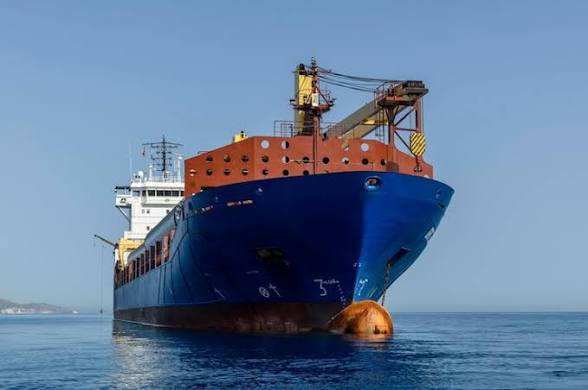 A large blue and rust-colored cargo ship floats on calm blue waters under a clear sky, viewed from the front showing its bow and crane structures.
