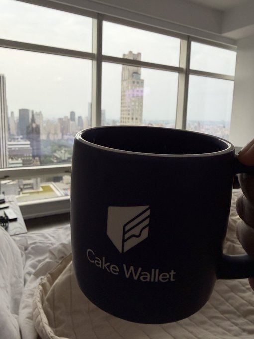A black 'Cake Wallet' branded coffee mug held up in front of a large window overlooking a city skyline with tall buildings.