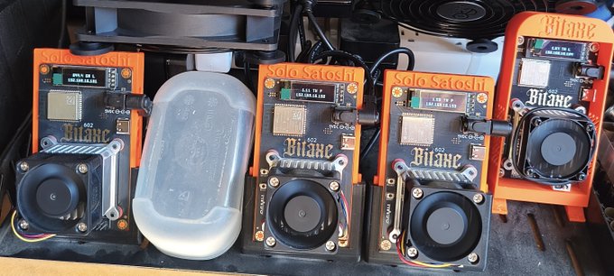 A collection of four BitAxe 'Solo Satoshi' cryptocurrency mining devices with orange cases and cooling fans, arranged on a shelf alongside a clear plastic container.