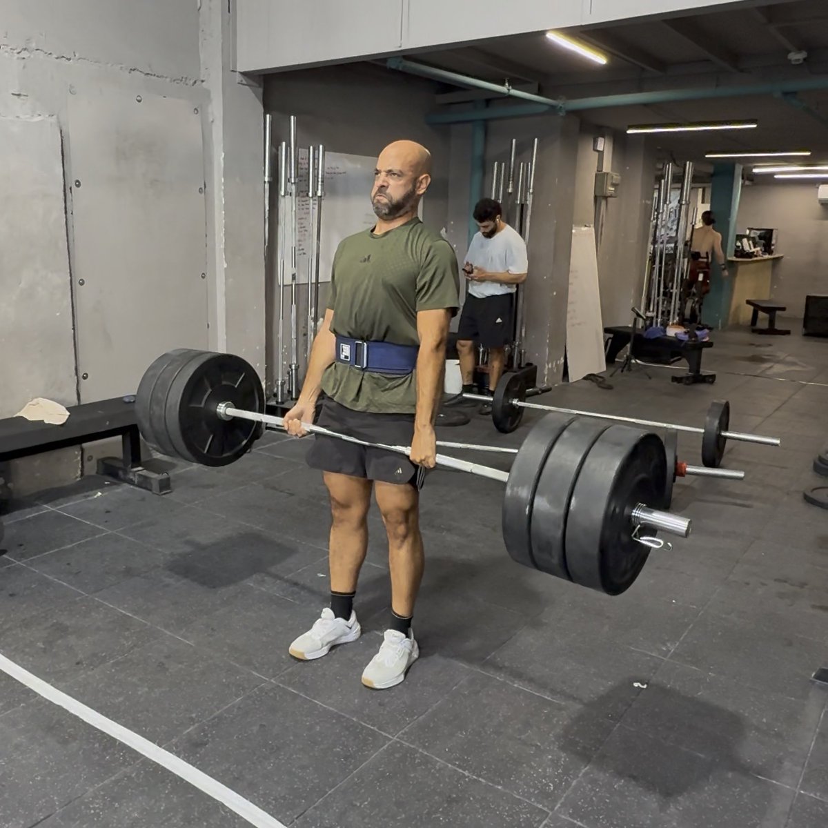 A bald, bearded man in an olive green t-shirt and a lifting belt performs a deadlift with a heavily loaded barbell in an industrial-style gym, with another man in a white shirt visible in the background.