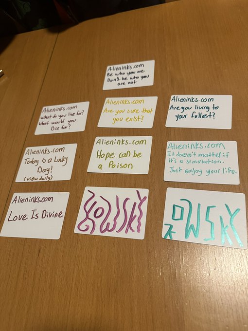 A spread of eight small white cards on a wooden table; the website 'Alieninks.com' is written on each, with various phrases like 'Be who you are', 'Hope can be a Poison', and 'Love Is Divine', alongside two cards with alien-like symbols.