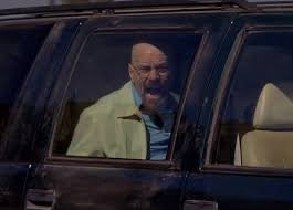 An older man with glasses and a green shirt sitting in the back seat of a car, making an exaggerated facial expression with his mouth open.