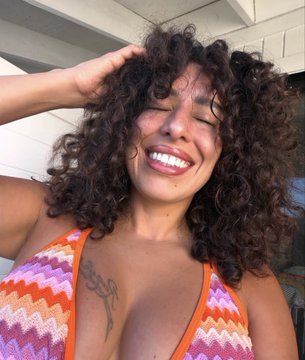 A woman with curly brown hair smiles and laughs while running her hand through her hair, wearing a colorful crochet top in an outdoor setting.