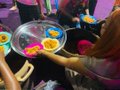 Children playing with colorful slime or paint on a clear circular table, with someone wearing red and black clothing visible.