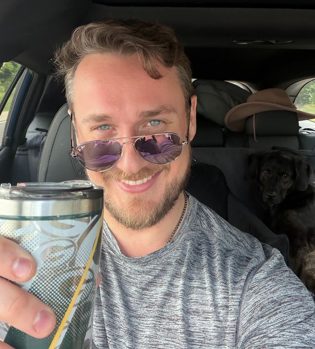 A smiling man wearing sunglasses takes a selfie in a car while holding a tumbler, with a dog visible in the back seat.