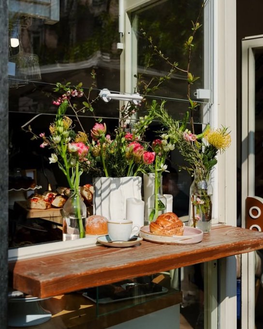 A cozy café scene featuring a wooden counter displaying glass vases filled with pink and yellow flowers, alongside pastries, a coffee cup, and a croissant.