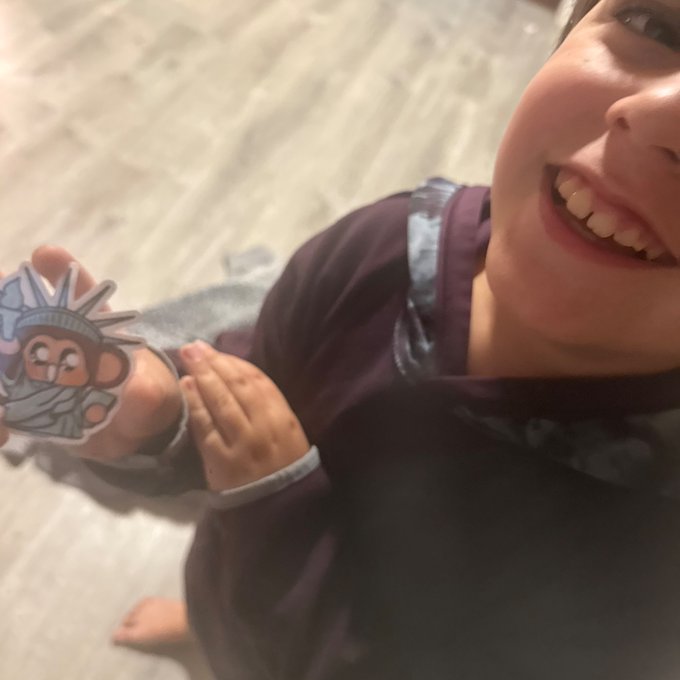 A close-up, slightly blurry selfie of a young girl smiling excitedly and holding up a cartoon sticker of a chibi Statue of Liberty.