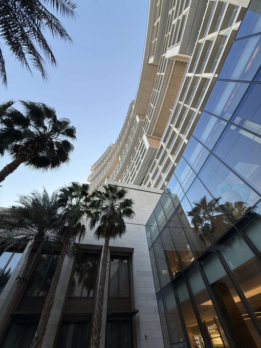 A low-angle shot captures a modern curved glass-front high-rise building against a clear blue sky, framed by tall palm trees in the foreground. The polished windows reflect the palms and sky, and a "BURBERRY" sign is partially visible at street level, suggesting the photo was taken outside a luxury retail destination. The upbeat "GM GM GM" caption likely nods to the sleek, glamorous aesthetic of the location, evoking a confident morning mood suited to a high-end shopping setting.
