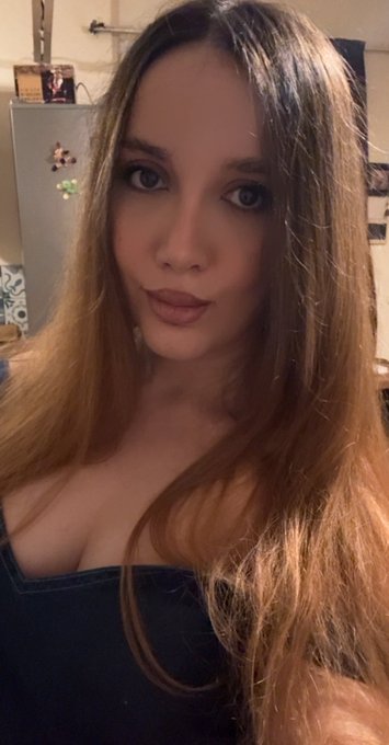 A selfie of a young woman with long brown hair wearing a dark top in a room with a refrigerator visible in the background.