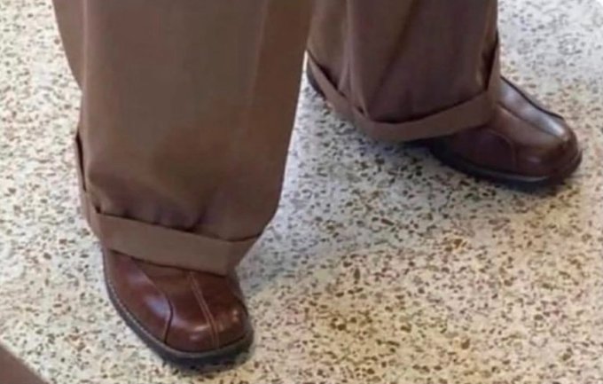 Close-up of a person's feet wearing brown leather shoes and tan pants with cuffed hems, standing on a speckled granite floor.