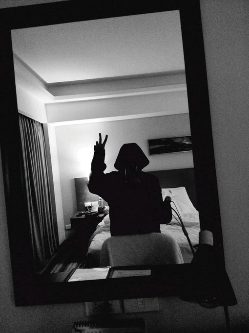 The image shows a person sitting at a desk with their hands raised, possibly making a peace sign or giving a thumbs up. The reflection of this scene can be seen in a mirror located near a bed. The background appears to be a bedroom setting, which includes a bed and a nightstand. There is also an open book on the nightstand next to the person's hand.