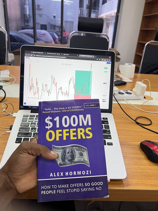The image shows a person holding up a book titled "100m Offers" over a laptop computer, possibly showing off some content or information from the book. The laptop displays a stock chart, which appears to be related to Bitcoin or Ethereum trading. Additionally, there are other people in the scene who seem to be watching something on a TV screen above their heads. This suggests that they might be following a live event or discussing financial markets with each other while observing some content displayed on the TV.