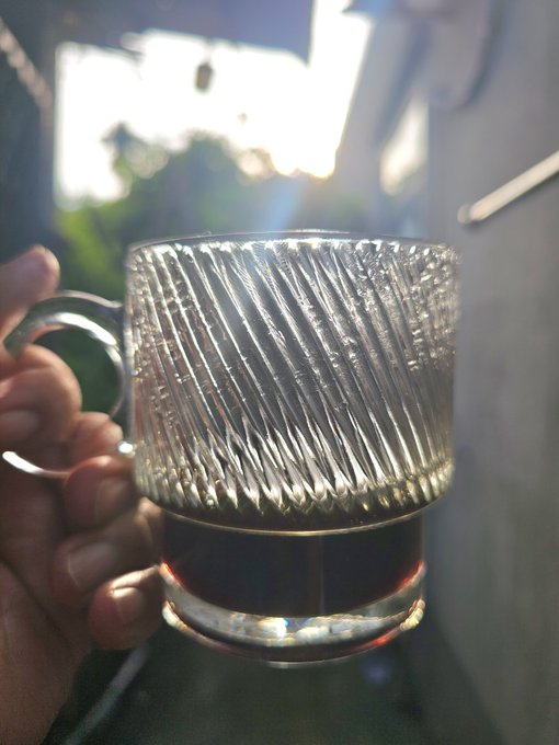 A hand holds a ribbed glass mug containing a small amount of dark liquid (likely coffee) up to the sunlight, creating a glowing effect through the textured glass.