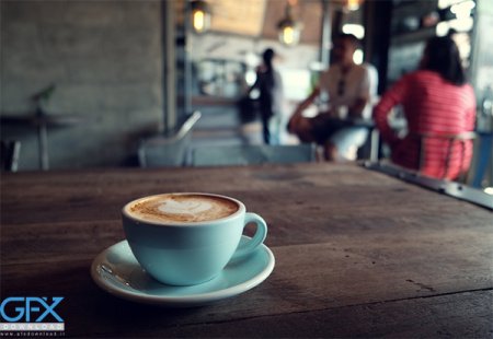 A light blue cup of cappuccino with heart latte art on a wooden table in a cafe, with blurred people in the background. The watermark 'GFX' appears in the bottom left corner.