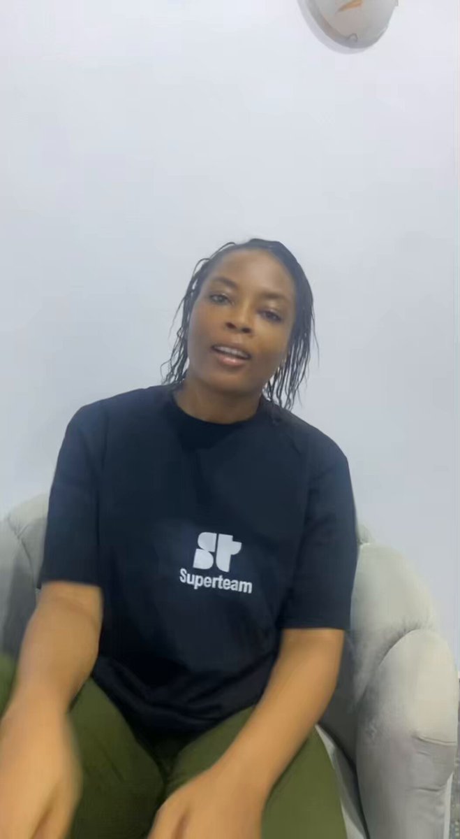 The image features a woman wearing a black shirt with white text, sitting down and smiling towards the camera. The background of the image is a plain white wall. There are no other objects or people visible in the image. The focus on the woman's face and her expression suggests that she might be participating in an online event or webinar related to the @SuperteamDE summit happening on Solana, which could be taking place at Spreespeicher.
