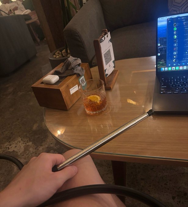 A hand holds a metallic vape pen in the foreground, with a wooden table behind it displaying a laptop, a glass with a drink and orange slice, and a wooden tissue box in a lounge setting.