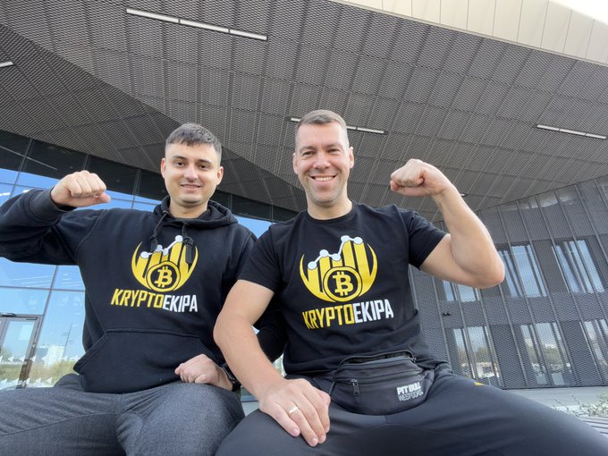 Two men wearing matching 'KRYPTOEKIPA' branded black apparel featuring a Bitcoin logo, posing confidently outside a modern building.