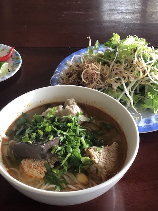 The image features two bowls of food sitting side by side on a wooden table. The bowl with soup contains noodles and vegetables, while the other bowl has salad with greens and sprouts. Both dishes are placed on blue plates that match each other. A small plate is also present in the scene, adding to the variety of food items displayed.