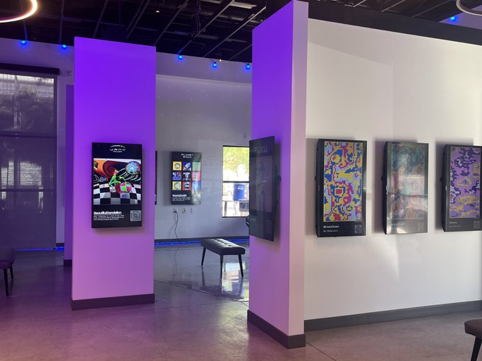 An exhibition space with purple-lit pillars displaying framed digital artwork and posters, suggesting an NFT or digital art gallery.