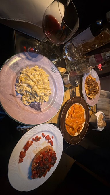 Overhead view of a dimly lit restaurant table featuring plates of pasta, bruschetta, and what appears to be a pumpkin dish, accompanied by a wine glass with red wine and a bottle.