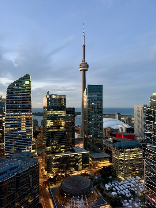 An aerial view of downtown Toronto at dusk, featuring the CN Tower, modern glass skyscrapers, and Lake Ontario in the background.