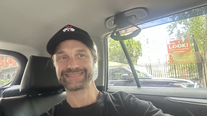 A selfie of a man wearing a black cap and black shirt, smiling inside a car, with a poster visible through the window that reads 'LOOK! FOR CYCLISTS'.
