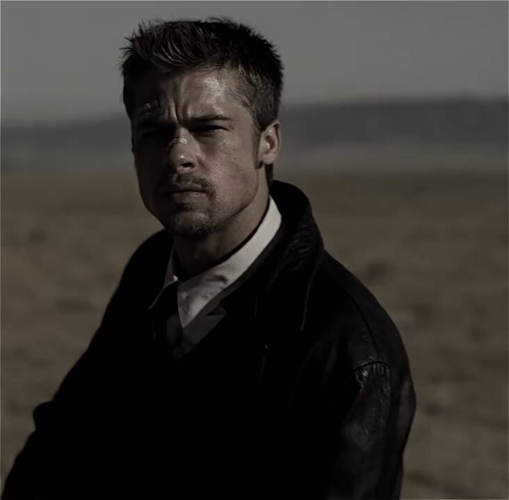 The image features a man wearing a suit jacket and tie, standing alone in an open field with his head tilted back towards the camera. The man appears to be looking directly at the viewer, creating a sense of connection between them. The background is a plain white wall that contrasts with the man's dark attire. There are no other people or objects visible in the image, which adds to its solitary atmosphere.