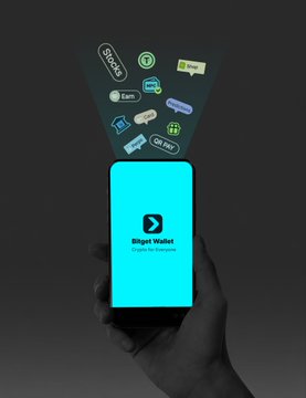 A hand holds a smartphone displaying the "Bitget Wallet" app logo on a teal screen, with various colorful crypto and NFT icons floating above the device, visually representing the wallet's enhanced features in cryptocurrency management, transactions, and Web3 security as highlighted in the post.