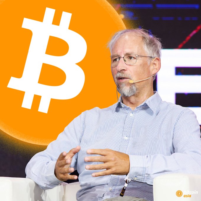 A man with glasses and a beard speaking at a conference, with a large Bitcoin logo displayed behind him.