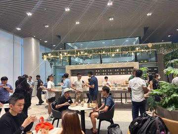 The image shows a group of people gathered around tables at an indoor restaurant or cafe. They are enjoying their time together while eating food and drinking from cups. The atmosphere appears lively as they engage in conversation, creating a sense of community and camaraderie among them. The scene is set against the backdrop of a large window that allows natural light to illuminate the space, enhancing the overall ambiance.