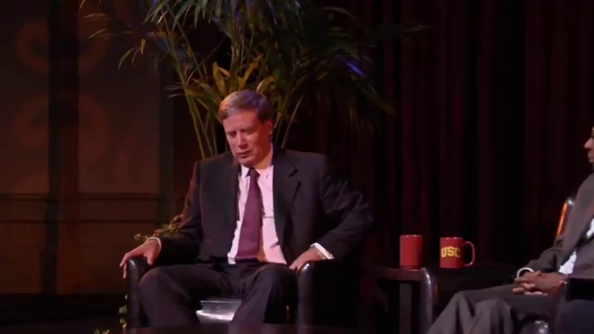 The image shows a man sitting in a chair, wearing a suit and tie, with a plant behind him. The man appears to be deep in thought or contemplation as he looks at something off-camera. In the background, there are two other people present, one of whom is holding a cup. The scene suggests that they might be attending an event or gathering where Trump has made some comments about the stock market and its performance over the past few days.