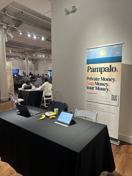 The image shows an office setting with a black table featuring two laptops, one of which has a banana peel sitting on top of it. The table also has a banner that says "Pampalodotcom" written on it. In the background, there are people working at tables and chairs scattered throughout the room. A sign is visible in the image as well.