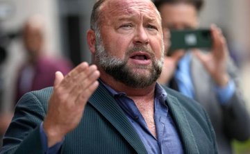 A close-up of a bearded man in a suit speaking, with photographers holding cameras visible in the background.