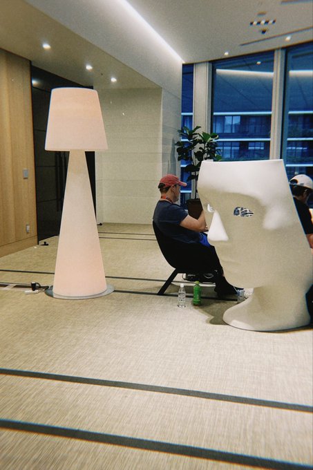 A man wearing a red cap sits in a chair next to a large white head sculpture in a modern office space, illuminated by a tall floor lamp.